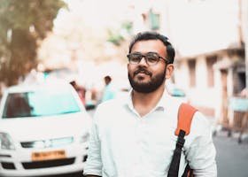 Confident man with glasses and backpack on a sunny urban street with cars in the background.