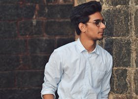 A stylish young man in glasses leans against a brick wall wearing a casual light blue shirt.